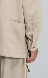 Camisa de manga larga color beige, estilo técnico con calce oversize. Presenta cuello camisero, cierre frontal de botones y múltiples bolsillos de parche con solapa en el frente, algunos con tiradores negros.