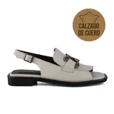 Sandalia de cuero color hueso con diseño tipo mocasín y detalle de hebillas metálicas. Tiene tira trasera con hebilla ajustable y suela plana color negro.