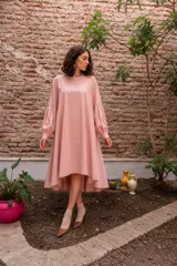 Vestido midi de algodón en color rosa empolvado, con silueta amplia y mangas largas voluminosas con detalle de tablas en los puños. Presenta un diseño de corte asimétrico, más corto en la parte frontal y más largo en la parte posterior.