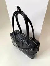 Cartera rígida negra con detalles en rojo y tachas plateadas. Tiene herrajes y cierre metálico plateado marca YKK, asa fina para llevar al hombro o en la mano y correa larga extra regulable. El interior es forrado, tiene un bolsillo con cierre enmarcado en cuero, y un segundo bolsillo plaqué.