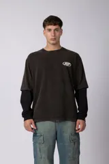 Remera de doble capa con diseño de efecto superpuesto. La capa exterior es de algodón con lavado acid wash en color gris oscuro y cuenta con un logo ovalado estampado en el pecho. La capa interior es de manga larga en color negro liso. Presenta un corte relajado y cuello redondo.