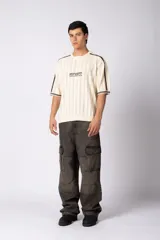 Remera de tejido de punto con textura calada, color crema con rayas verticales sutiles. Presenta cuello en V, mangas cortas con franjas laterales en color marrón oscuro y logo bordado en el centro del pecho.