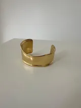 Brazalete rígido tipo cuff de diseño minimalista, acabado en tono dorado brillante.