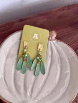 Aros colgantes con base rectangular texturizada dorada, de la cual cuelgan abalorios de cristal facetado en tonos verdes y pequeños detalles color salmón, junto a tres dijes colgantes en forma de pétalo u hoja de color verde.