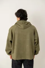 Canguro con capucha de estilo urbano en color verde oliva, confeccionado en tejido suave. Presenta un panel frontal en contraste de color gris oscuro con cierre de cremallera parcial, mangas largas con puños elásticos y bolsillos laterales integrados.