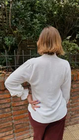 Camisa blanca de textura sutil, con cuello alto tipo victoriano y detalle de volados asimétricos a lo largo de la botonadura frontal. Presenta mangas largas con puños terminados en volados.