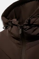 Cazadora acolchada color marrón con capucha ajustable, cuello subido y cierre frontal de cremallera. Presenta diseño combinado con mangas largas de tejido liso y cuerpo acolchado con costuras en chevrón, además de bolsillos laterales con cierre invisible.
