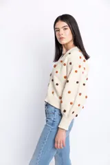 Cárdigan de lana color crudo con pompones marrones y naranjas, escote en V, cierre frontal con botones de madera, mangas largas abullonadas y corte holgado.