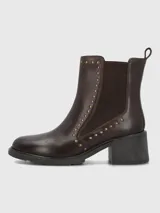 Bota tipo botín de cuero color marrón oscuro, con elásticos laterales y detalles de tachas metálicas doradas a lo largo de la caña. Cuenta con suela gruesa y taco cuadrado de altura media.