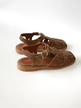 Sandalias estilo cangrejera color marrón, confeccionadas en cuero con diseño trenzado en la parte delantera, tira ajustable con hebilla metálica en el tobillo y suela plana.