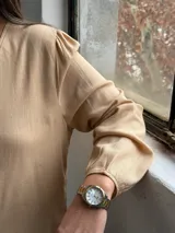 Blusa de color tostado con cuello en V, cierre frontal con botones y mangas largas con puños elásticos. Presenta un diseño de corte recto con bolsillos laterales integrados.