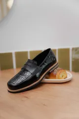 Mocasines negros de cuero con textura tipo cocodrilo en la parte superior y acabado brillante. Presentan una banda frontal clásica y una suela plana con detalle de borde en color natural.