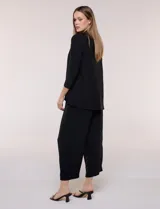 Cardigan negro de tejido elastizado con textura lisa, frente abierto sin botones y solapas amplias de caída drapeada. Cuenta con mangas tres cuartos y ruedo asimétrico.
