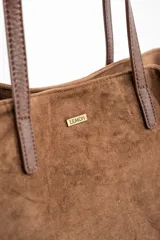 Bolso tipo tote de gran tamaño confeccionado en gamuza sintética color tostado. Presenta un diseño minimalista con asas largas de hombro y una pequeña placa metálica frontal con el logo de la marca.