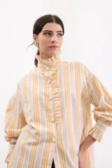 Camisa holgada de algodón con estampado de rayas en tonos amarillos, naranjas y grises. Presenta cuello alto con volados, cierre frontal con botones dorados y mangas largas abullonadas con puños con volados.