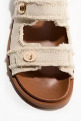 Sandalias planas de sarga de algodón en color beige con terminación desflecada. Cuentan con tiras ajustables con detalle metálico dorado y cierre autoadherente, además de una tira ajustable en el talón. La plantilla es preformada en tono marrón.