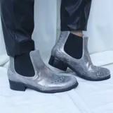 Botín tipo Chelsea de cuero metalizado en tono bronce, con paneles elásticos laterales en color beige. Presenta detalles de costuras decorativas tipo brogue en la puntera y laterales, con suela de goma antideslizante y taco bajo foliado.