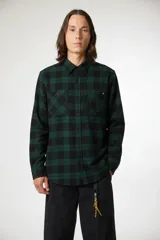 Camisa de manga larga con estampado de cuadros en tonos verde y negro, confeccionada en tela tipo franela. Presenta cuello clásico, cierre frontal con botones y dos bolsillos frontales en el pecho.