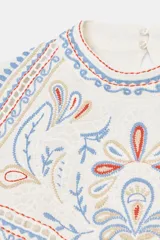 Camisa de cuello redondo y manga corta con diseño holgado. Presenta bordados a contraste en tonos azules, rojos y beige en la parte superior delantera y hombros. Cuenta con cierre posterior mediante abertura y botones.