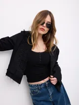 Chaqueta corta negra de gabardina con detalles de cordones decorativos bordados en el frente y bordes deshilachados.