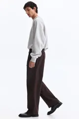 Pantalón sastrero color marrón oscuro, de corte relajado y piernas con pliegues, confeccionado en mezcla de algodón y lyocell. Cuenta con cierre y botón, bolsillos laterales y bolsillos traseros insertados con botón oculto.