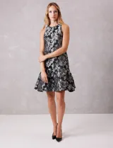Vestido corto negro con estampado floral en jacquard metalizado plateado. Presenta escote a la base sin mangas, corte recto en el cuerpo y un gran volado en el ruedo. Incluye cierre invisible en la espalda y forro interno.