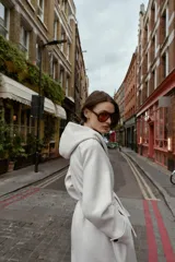 Abrigo de paño negro con capucha, cierre frontal cruzado con botones y cinturón de lazada en el mismo tejido. Presenta manga larga con trabillas en los puños y bolsillos laterales.