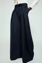 Pantalón de tiro medio de corte wide leg, confeccionado en mezcla de lyocell con patrón de rayas diplomáticas. Presenta bolsillos laterales ocultos, pliegues delanteros marcados y cierre frontal con cremallera, botón y ganchos metálicos.