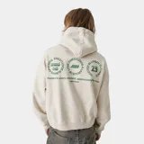 Hoodie de corte oversized color beige con capucha y bolsillo frontal tipo canguro. Presenta estampados gráficos circulares en color verde en la espalda con tipografía y logos de la marca.