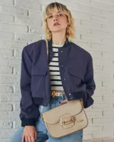 Cartera negra de cuero charolado con solapa y cierre de broche. Presenta un detalle metálico dorado en forma de freno de caballo en la parte frontal y una correa ajustable.