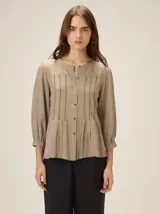 Camisa de lino mezcla color beige, con cuello redondo, cierre frontal con botones y mangas tres cuartos con puños elásticos. Presenta detalles de pespuntes verticales en zigzag en el frente.