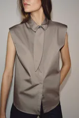 Camisa sin mangas confeccionada en algodón, con cuello redondo y detalle de lazada frontal. Presenta cierre frontal con botones ocultos por solapa y bajo redondeado.