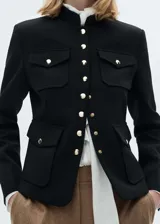 Chaqueta de estilo militar en color negro, con cuello redondo, manga larga y cierre frontal de botones metálicos dorados. Presenta cuatro bolsillos de solapa en la parte delantera y diseño de corte recto.