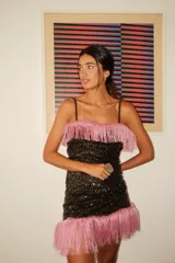 Vestido corto sin mangas, color negro, completamente cubierto de lentejuelas brillantes. Presenta un borde de flecos largos de color rosado en el escote y en el ruedo inferior.