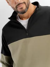 Buzo de felpa con diseño color block, combinando negro en la parte superior y beige en la parte inferior. Presenta cuello alto con cierre frontal parcial y puños elásticos.