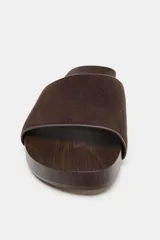 Sandalia plana con tira ancha de gamuza marrón y suela con efecto madera. Presenta detalle de tachas metálicas en el lateral y punta redonda.