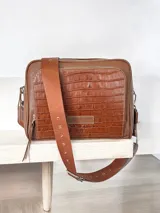 Bandolera marrón de cuero genuino con textura tipo cocodrilo en el frente. Tiene correa de tela con el nombre de la marca y herrajes plateados.