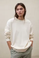 Buzo de cuello redondo color blanco marfil, con hombros caídos y diseño oversize. Cuenta con puños, cuello y bajo acanalados, además de detalles de costura decorativa en la parte frontal del cuello.