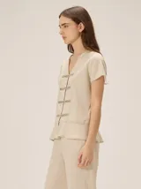 Camisa de manga corta color beige con estampado a cuadros, cuello en V y detalles de galones decorativos estilo oriental en la parte frontal.