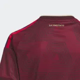 Camiseta de fútbol Adidas, modelo Local 2026 para fanáticos, color granate con patrón geométrico de triángulos en tono más oscuro. Presenta cuello en V con ribete beige y detalles en amarillo y azul en los hombros. Lleva el escudo de la selección venezolana y el logo de Adidas bordados.
