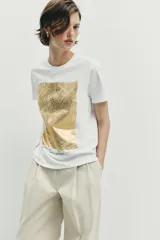Camiseta de algodón con cuello redondo y manga corta. Presenta un diseño de estampación metalizada en color dorado en la parte frontal con detalles de texto.
