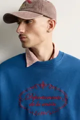 Sudadera azul con corte relaxed fit, cuello redondo y manga larga, confeccionada en tejido de algodón. Presenta un bordado frontal a contraste en color rojo con diseño ornamental y texto.