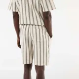 Bermuda de corte clásico confeccionada en tejido ligero de color beige con diseño de rayas verticales oscuras. Cuenta con bolsillos laterales y pretina con elástico.
