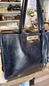 Cartera tipo tote de cuero negro con acabado texturizado, cierre frontal giratorio metálico en tono dorado y detalle de borla lateral. Cuenta con doble asa de hombro.