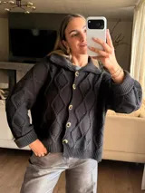 Saco tejido en color verde oliva con diseño de ochos y trenzas. Presenta cuello estilo camisero, cierre frontal con botones tipo joya, mangas holgadas y silueta oversize.