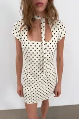 Vestido corto color crema con estampado de lunares negros. Presenta cuello redondo con lazada en la parte trasera, escote cuadrado, manga corta y cierre de cremallera oculta en la espalda.