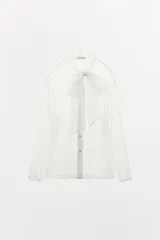Camisa semitransparente de organza en color blanco, con cuello subido y lazada frontal. Presenta manga larga con puños abotonados y cierre frontal con botones ocultos.