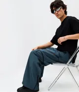 Pantalón de corte holgado en color azul petróleo, confeccionado en microfibra de poliéster. Presenta un diseño utilitario con múltiples bolsillos funcionales, cierres visibles y detalles técnicos en las piernas.