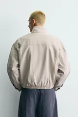 Cazadora relaxed fit confeccionada en tejido de algodón de color beige, con cuello subido y manga larga. Cuenta con bolsillos de plastrón en la cadera, bajo elástico y cierre frontal con cremallera oculta y alamares metálicos.