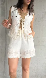 Conjunto de chaleco y short de lino color natural con bordados florales. El chaleco se ajusta con lazo frontal y el short tiene ruedo festoneado.